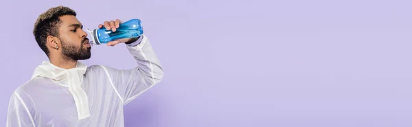 Bearded african american sportsman holding sports bottle and drinking refreshing water on purple, banner — Stock Photo