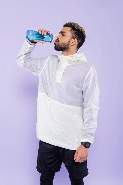 Junger afrikanisch-amerikanischer Sportler hält Sportflasche in der Hand und trinkt erfrischendes Wasser auf violett — Stockfoto