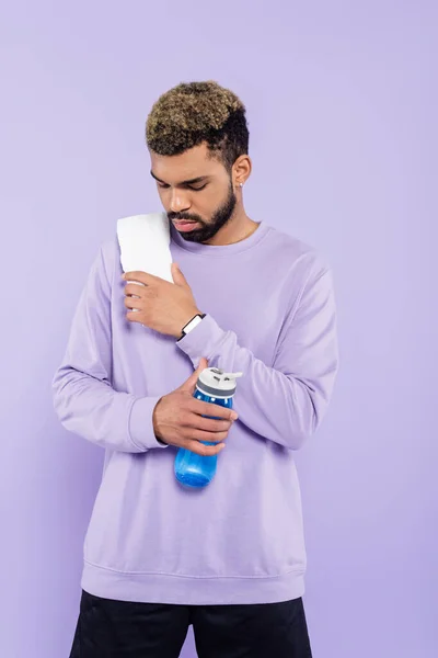 Uomo afroamericano barbuto in maglione con asciugamano bianco in possesso di bottiglia sportiva con acqua isolata su viola — Foto stock