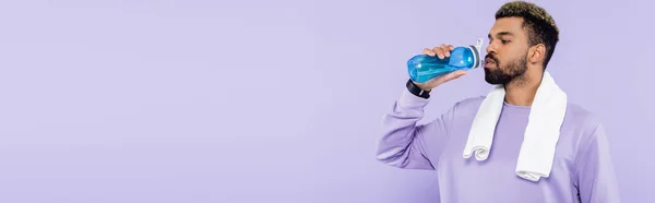 Bearded african american man in sweater standing with white towel and drinking water isolated on purple, banner — Stock Photo