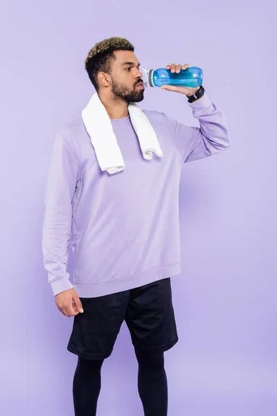 Bearded african american man in sweater standing with white towel and drinking water isolated on purple — Stock Photo