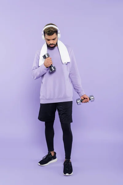 Pleine longueur de barbu homme afro-américain dans les écouteurs exercice avec haltères sur violet — Photo de stock