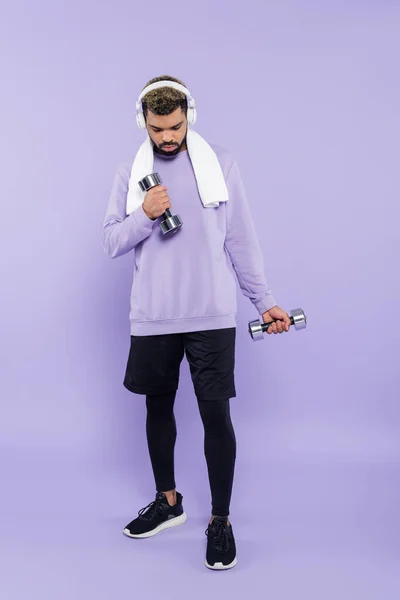 Comprimento total do homem americano africano barbudo em fones de ouvido exercitando com halteres em roxo — Fotografia de Stock