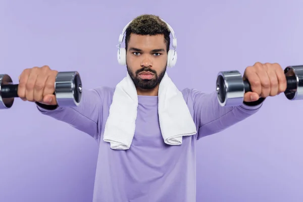 Young african american man in headphones exercising with dumbbells isolated on purple — Stock Photo