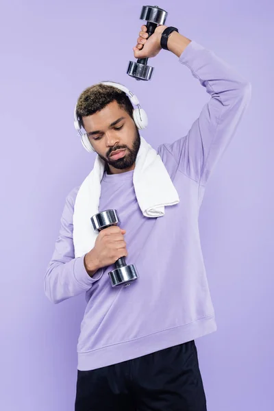 Homme afro-américain barbu dans des écouteurs sans fil exercice avec haltères isolés sur violet — Photo de stock