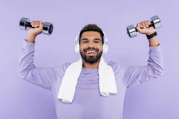 Heureux homme afro-américain dans les écouteurs exercice avec haltères isolé sur violet — Photo de stock