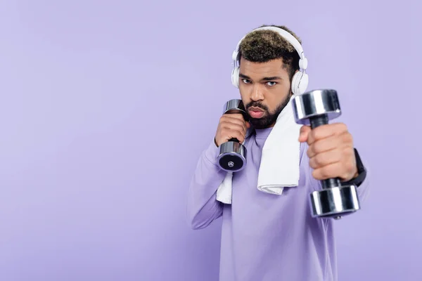 Esportivo afro-americano em fones de ouvido exercitando com halteres isolados em roxo — Fotografia de Stock