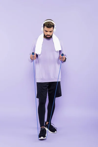 Full length of bearded african american man in wireless headphones holding jumping rope on purple — Stock Photo