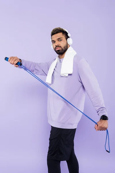 Bearded african american man in wireless headphones holding jumping rope isolated on purple — Stock Photo