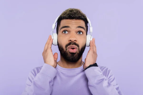 Hombre afroamericano sorprendido y barbudo en auriculares inalámbricos aislados en púrpura - foto de stock