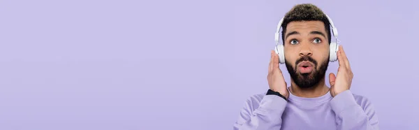 Surpreso e barbudo afro-americano em fones de ouvido sem fio isolados em roxo, banner — Fotografia de Stock