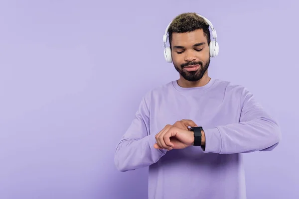 Homme afro-américain barbu en écouteurs sans fil regardant montre intelligente isolé sur violet — Photo de stock