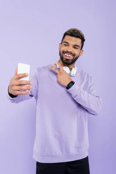 Glücklicher afrikanisch-amerikanischer Mann mit drahtlosen Kopfhörern, der ein Selfie macht, während er das Friedenszeichen isoliert auf lila zeigt — Stockfoto