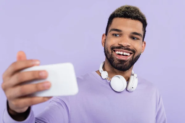 Homem americano africano alegre com fones de ouvido sem fio levando selfie isolado em roxo — Fotografia de Stock