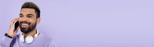 Hombre afroamericano feliz en auriculares inalámbricos hablando en el teléfono inteligente aislado en púrpura, bandera - foto de stock