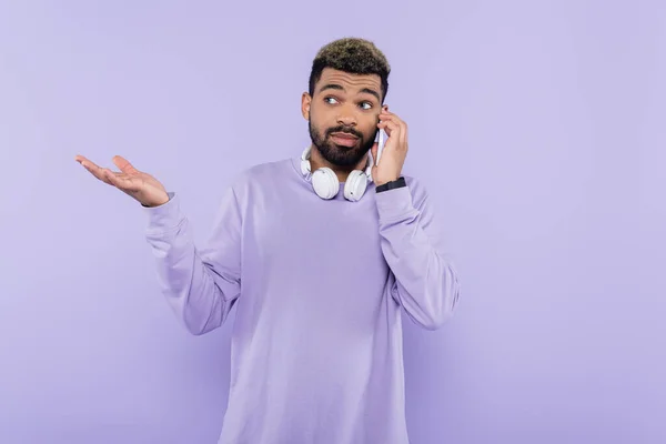 Bearded african american man with wireless headphones talking on smartphone isolated on purple — Stock Photo