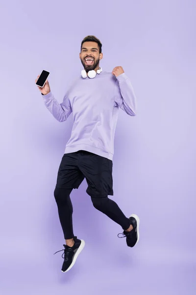 Збуджений афроамериканський чоловік з бездротовими навушниками, що тримає смартфон і стрибає на фіолетовому — стокове фото