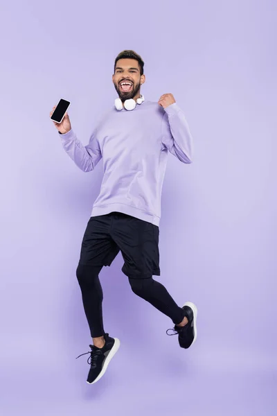 Animado afro-americano homem com fones de ouvido sem fio segurando smartphone e pulando em roxo — Fotografia de Stock