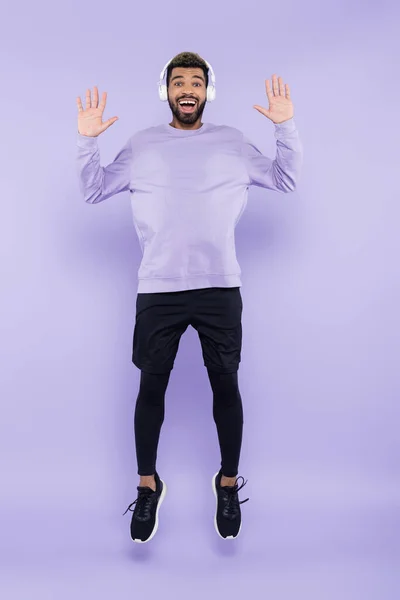 Pleine longueur de l'homme afro-américain excité dans les écouteurs sans fil sautant sur violet — Photo de stock