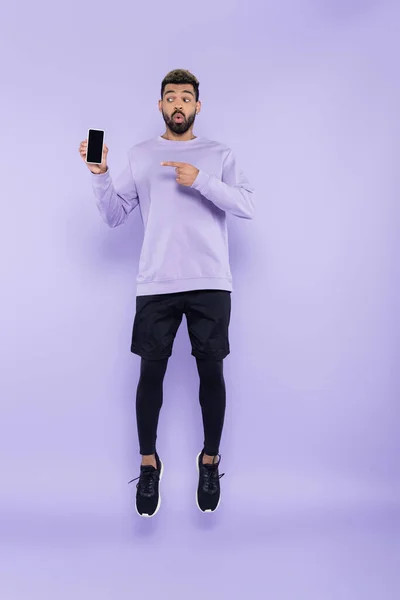Full length of surprised african american man pointing at smartphone and levitating on purple — Stock Photo