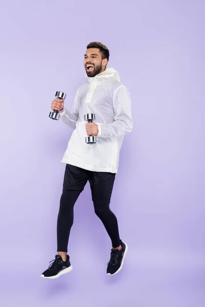 Full length of excited african american man in sportswear levitating and holding dumbbells on purple — Stock Photo