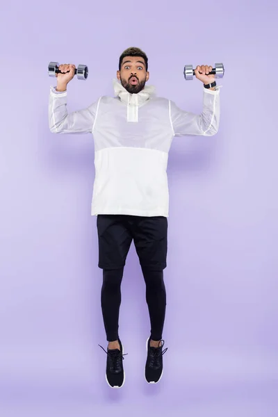 Full length of shocked african american man in sportswear levitating and holding dumbbells on purple — Stock Photo