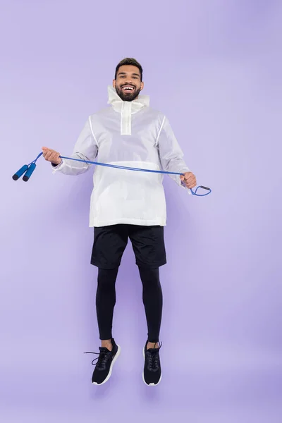 Pleine longueur de heureux homme afro-américain en vêtements de sport lévitant et tenant la corde sautante sur violet — Photo de stock