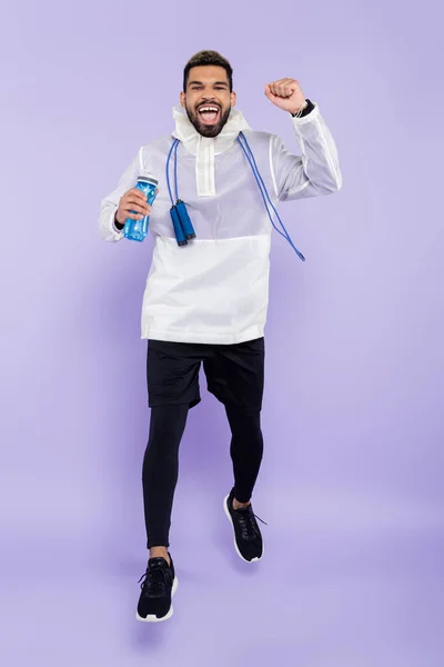 Comprimento total do homem americano africano animado em sportswear levitando e segurando garrafa de esportes em roxo — Fotografia de Stock