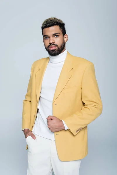 Hombre afroamericano con estilo en chaqueta amarilla posando con la mano en el bolsillo aislado en gris - foto de stock