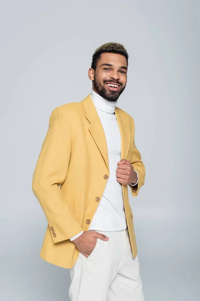 Hombre americano africano alegre en chaqueta amarilla posando con la mano en el bolsillo aislado en gris - foto de stock