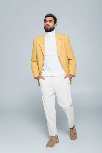 Comprimento total de homem americano africano elegante em blazer amarelo posando com as mãos em bolsos em cinza — Fotografia de Stock