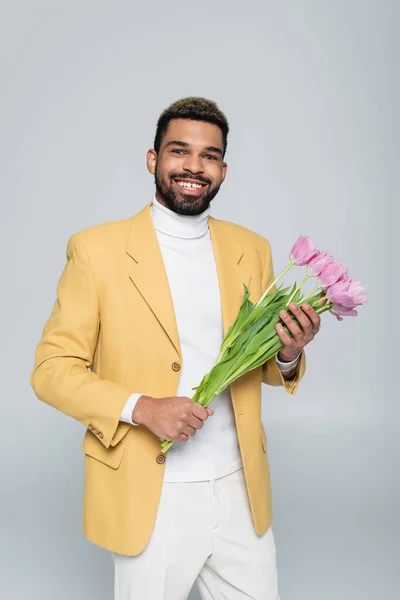 Felice uomo africano americano in abito elegante che tiene bouquet di tulipani rosa isolati su grigio — Foto stock