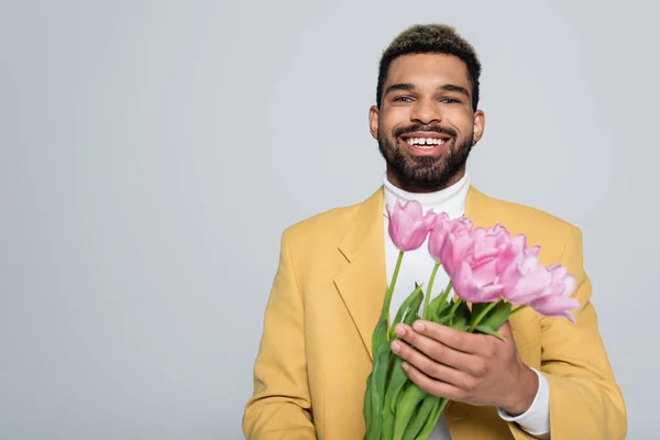 Allegro uomo afroamericano in abito elegante che tiene bouquet di tulipani rosa isolati su grigio — Foto stock