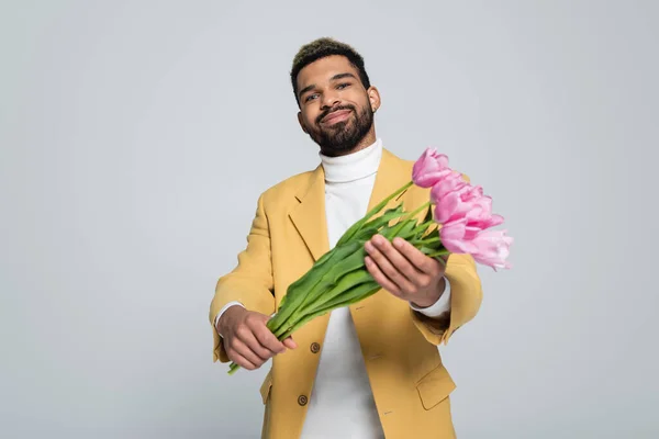 Hombre afroamericano complacido en traje elegante celebración ramo de tulipanes rosados aislados en gris - foto de stock