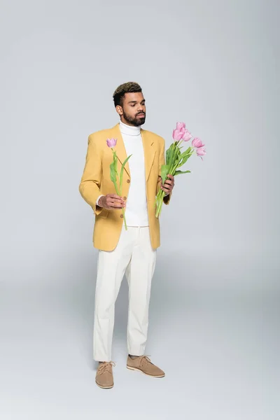 Longitud completa de hombre afroamericano en traje elegante celebración ramo de tulipanes rosados en gris - foto de stock