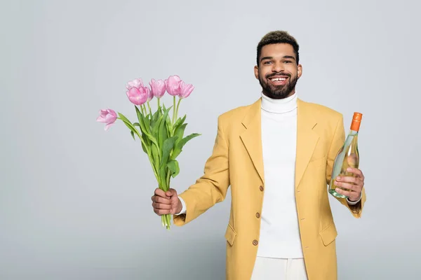 Happy african american man in stylish outfit holding bouquet of pink tulips isolated on grey — Stock Photo
