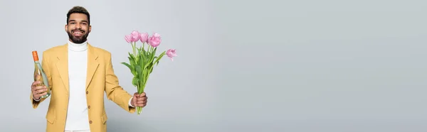 Positive african american man in stylish outfit holding bouquet of pink tulips isolated on grey, banner — Stock Photo