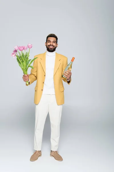 Full length of happy african american man in stylish outfit holding bouquet of tulips and bottle of champagne on grey — Stock Photo