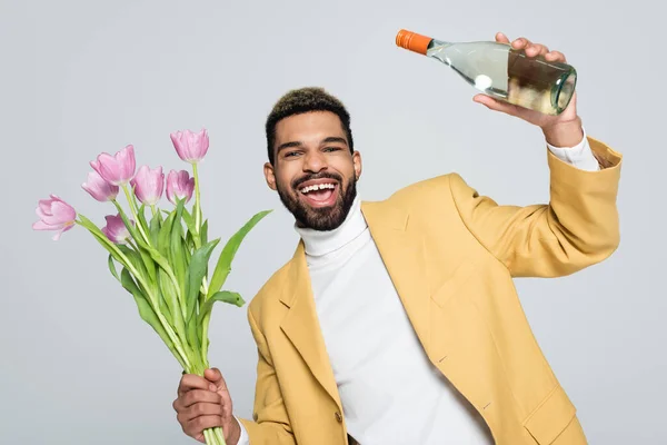 Excité homme afro-américain en tenue élégante tenant bouquet de tulipes roses et bouteille de champagne isolé sur gris — Photo de stock