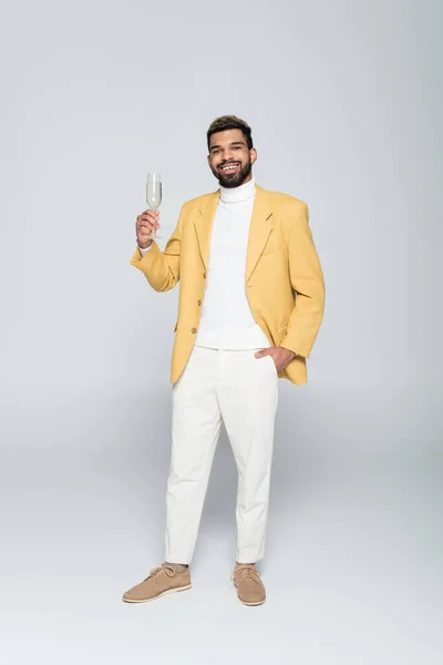 Volle Länge der fröhlichen afrikanisch-amerikanischen Mann in stilvollem Outfit hält ein Glas Champagner und posiert auf grau — Stockfoto