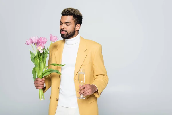 Jovem afro-americano em blazer amarelo e pescoço pólo segurando tulipas rosa e copo de champanhe isolado em cinza — Fotografia de Stock