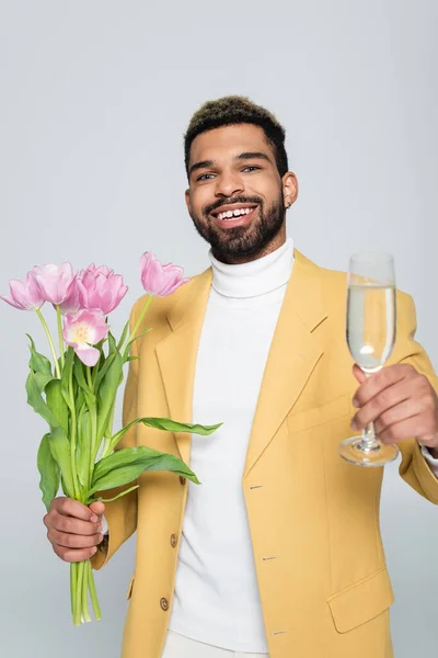 Happy african american man in yellow blazer and polo neck holding pink tulips and glass of champagne isolated on grey — Stock Photo