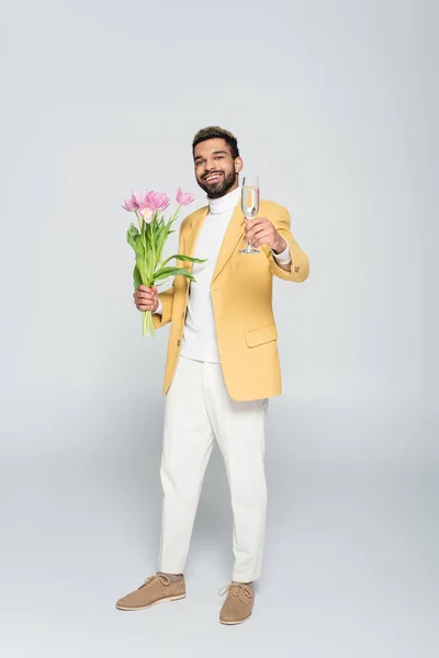 Longitud completa de hombre afroamericano feliz en chaqueta amarilla con tulipanes rosados y copa de champán aislado en gris - foto de stock