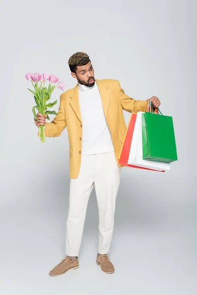 Sorprendido hombre afroamericano en blazer amarillo y cuello polo sosteniendo tulipanes rosados y bolsas de compras en gris - foto de stock