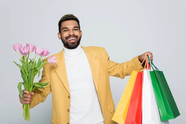 Homem americano africano alegre em blazer amarelo e pescoço pólo segurando tulipas rosa e sacos de compras isolados em cinza — Fotografia de Stock