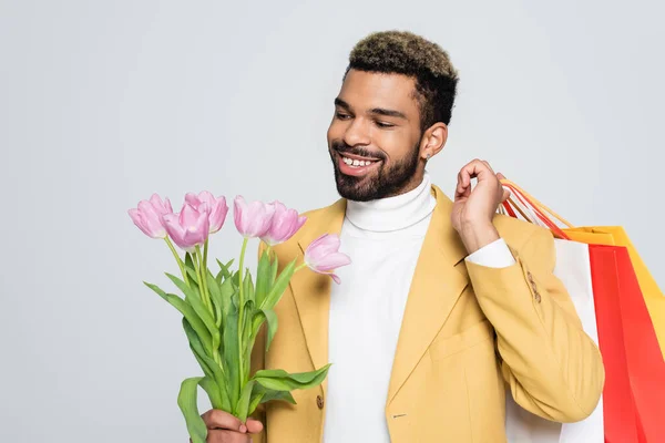 Homem americano africano alegre em blazer amarelo e pescoço pólo segurando tulipas rosa e sacos de compras isolados em cinza — Fotografia de Stock