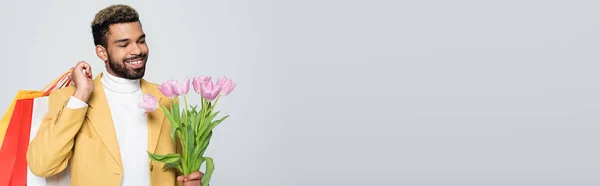 Felice uomo afroamericano in giacca gialla e collo polo con tulipani rosa e shopping bag isolati su grigio, banner — Foto stock
