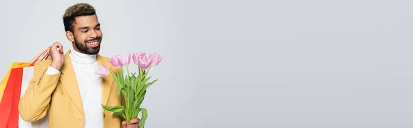 Hombre afroamericano feliz en blazer amarillo y cuello polo sosteniendo tulipanes rosados y bolsas de compras aisladas en gris, bandera - foto de stock