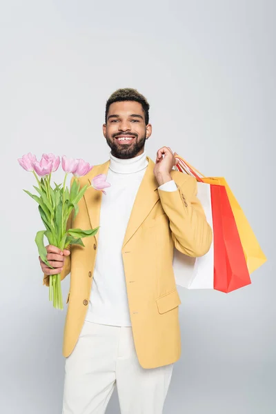 Positive african american man in yellow blazer and polo neck holding pink tulips and shopping bags isolated on grey — Stock Photo