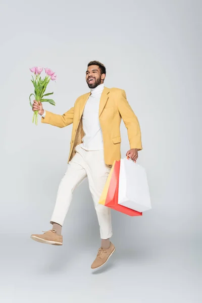 Pleine longueur de joyeux homme afro-américain en blazer jaune et col polo tenant des tulipes roses et des sacs à provisions sur gris — Photo de stock